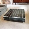 Table basse carrée avec plateau carrelé vert et joints blancs, placée dans un salon moderne avec canapé gris, tapis clair et meuble en bois.