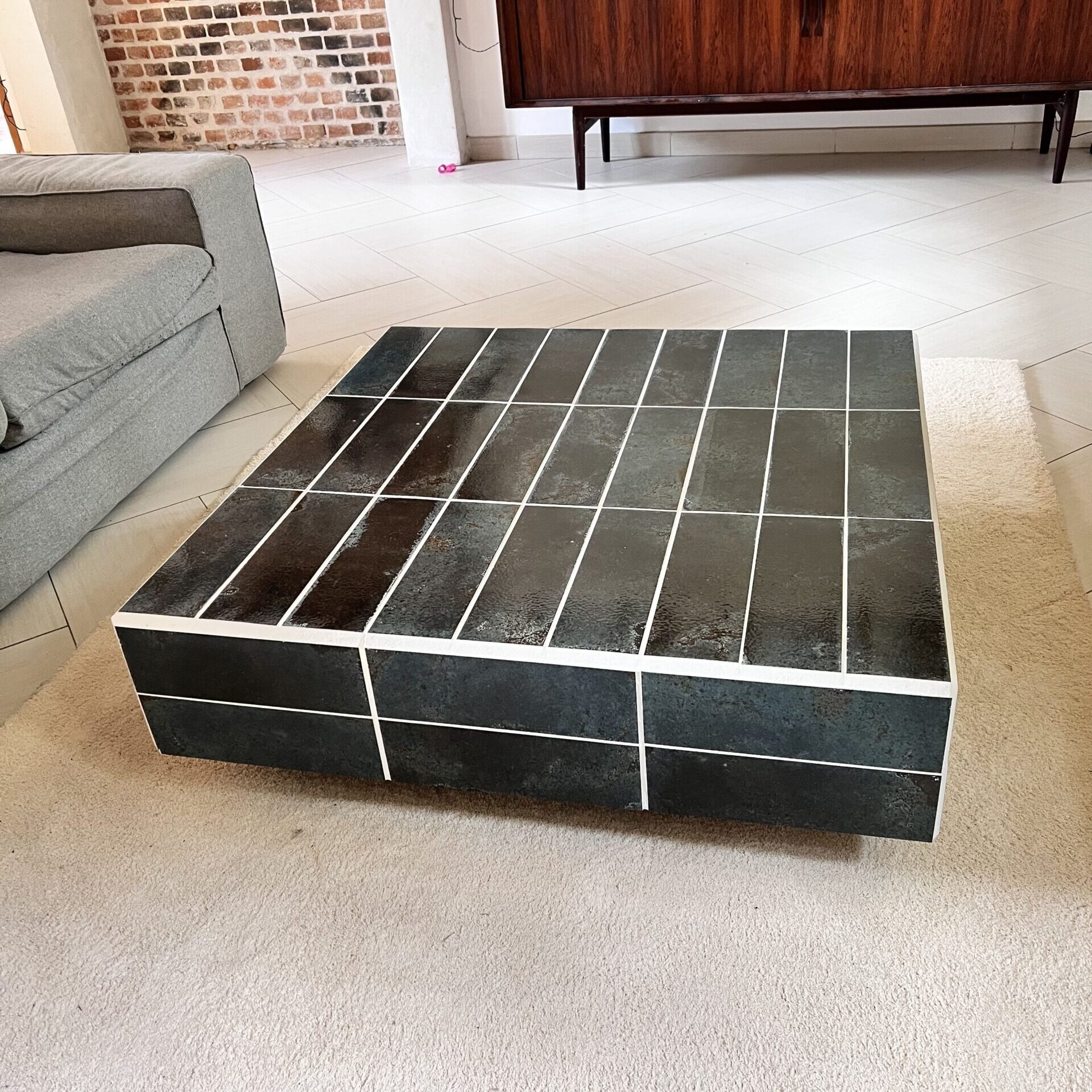 Table basse carrée avec plateau carrelé vert et joints blancs, placée dans un salon moderne avec canapé gris, tapis clair et meuble en bois.